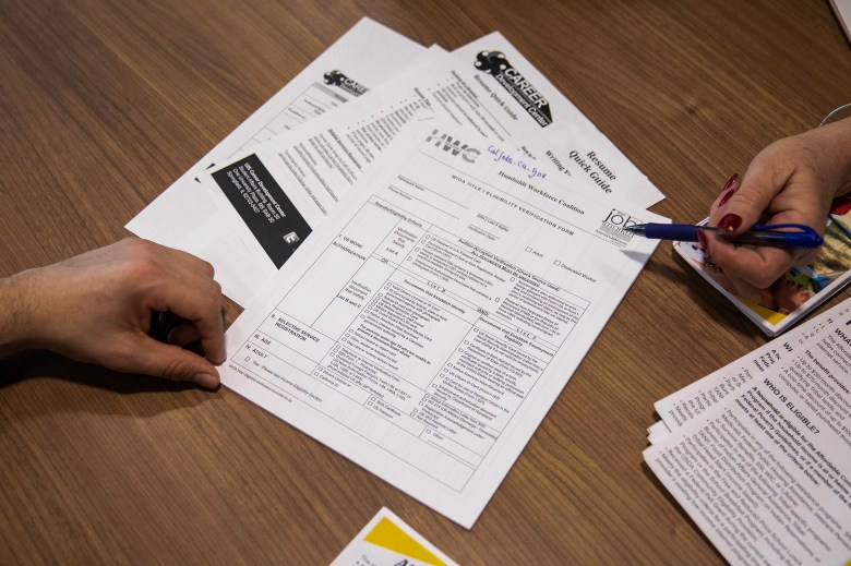 A small stack of papers sit on a brown wooden desk, while one hand holds an edge of a paper, and another points at an opposite corner with a blue pen. The setting is a job related workshop or fair. The words, "Career", "Resume" and "Workforce" are visible on the papers.