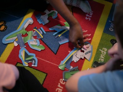 Close-up of several hands working on a colorful puzzle on a brightly patterned carpet. One hand, with neatly manicured nails, reaches for a puzzle piece, while another figure, partially visible, observes or assists. The carpet features vibrant red, blue, green, and yellow sections with printed text and designs. The atmosphere suggests collaborative play or learning.