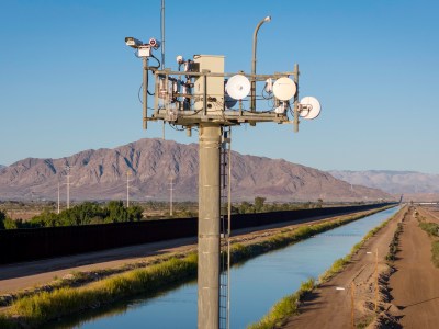 A surveillance tower monitor, with a background landscape of a brown fence, river and mountains.