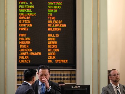 Two individuals are engaged in a private conversation in the foreground of a legislative chamber, with one appearing to whisper to the other. A digital display board in the background lists names in green, red, and orange, indicating legislative votes or attendance. A third person in a suit and glasses sits nearby, observing the scene. Bookshelves and muted green walls with gold accents frame the setting.