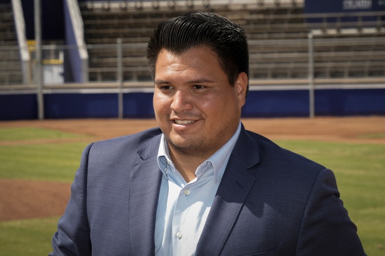 City of Compton councilman Isaac Galvan, wearing a blue suit smiles while standing in front of a baseball field, with empty stands in the background.