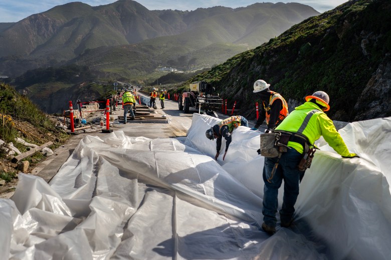 A group of construction workers in safety gear work on a roadside project near a steep, mountainous landscape. Large sheets of white plastic are being secured to the ground, with some workers bending to adjust the material while others stand nearby. Traffic cones, barriers, and equipment line the road, which is surrounded by lush green hills and rocky cliffs under a clear sky.