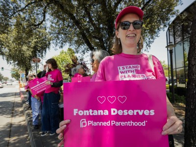 A group of individuals wearing bright pink Planned Parenthood shirts stand along a sidewalk under the shade of trees, holding signs in support of Planned Parenthood. In the foreground, one person wearing sunglasses and a pink cap smiles while holding a sign that reads "Fontana Deserves Planned Parenthood" with three heart icons above the text.