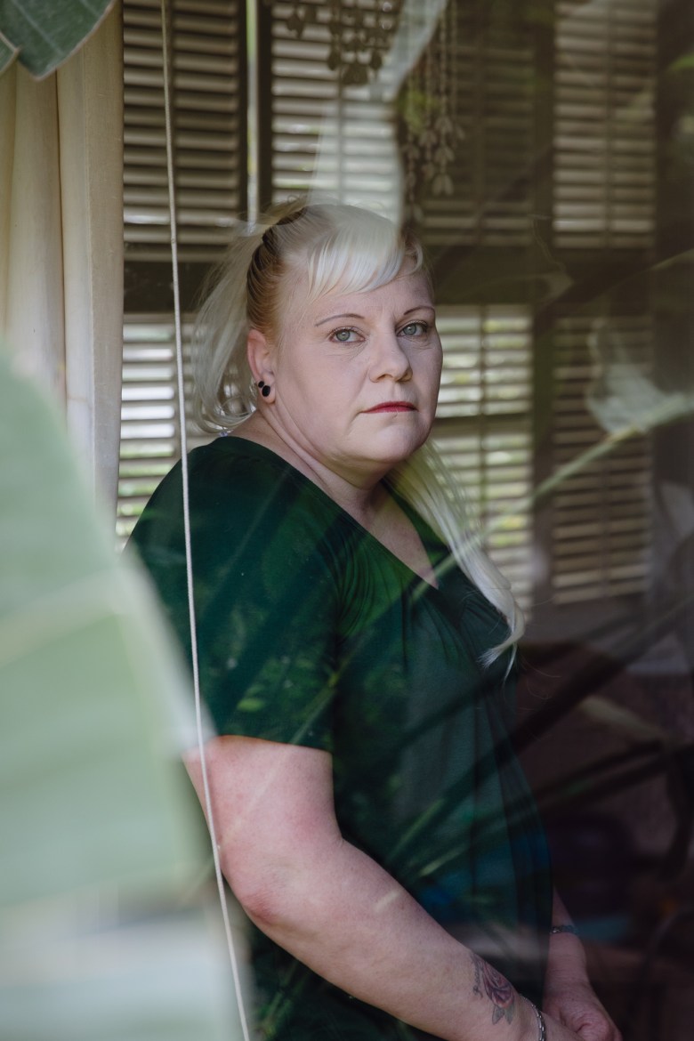 A woman sits in her home with a quiet face looking straight at the camera through a glass window. Some leaves from outside frame the left side of the image and the glass window reflects branches and leaves from outside.