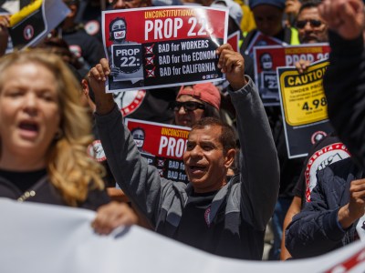 A crowd of protesters hold Prop. 22 signs with their mouths open and fists in the air. One prominent sign reads, "Unconstitutional Prop 22, Bad for Workers, Bad for the Economy, Bad for California."