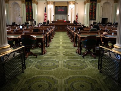 An empty legislative chamber featuring rows of dark wooden desks and green leather chairs arranged neatly on an ornate green carpet. Two large boards with illuminated text hang on either side of the speaker's podium at the front of the room. The American and Californian flags are positioned behind the podium, framed by tall, elegant columns. The quiet setting suggests a pause in legislative activity.