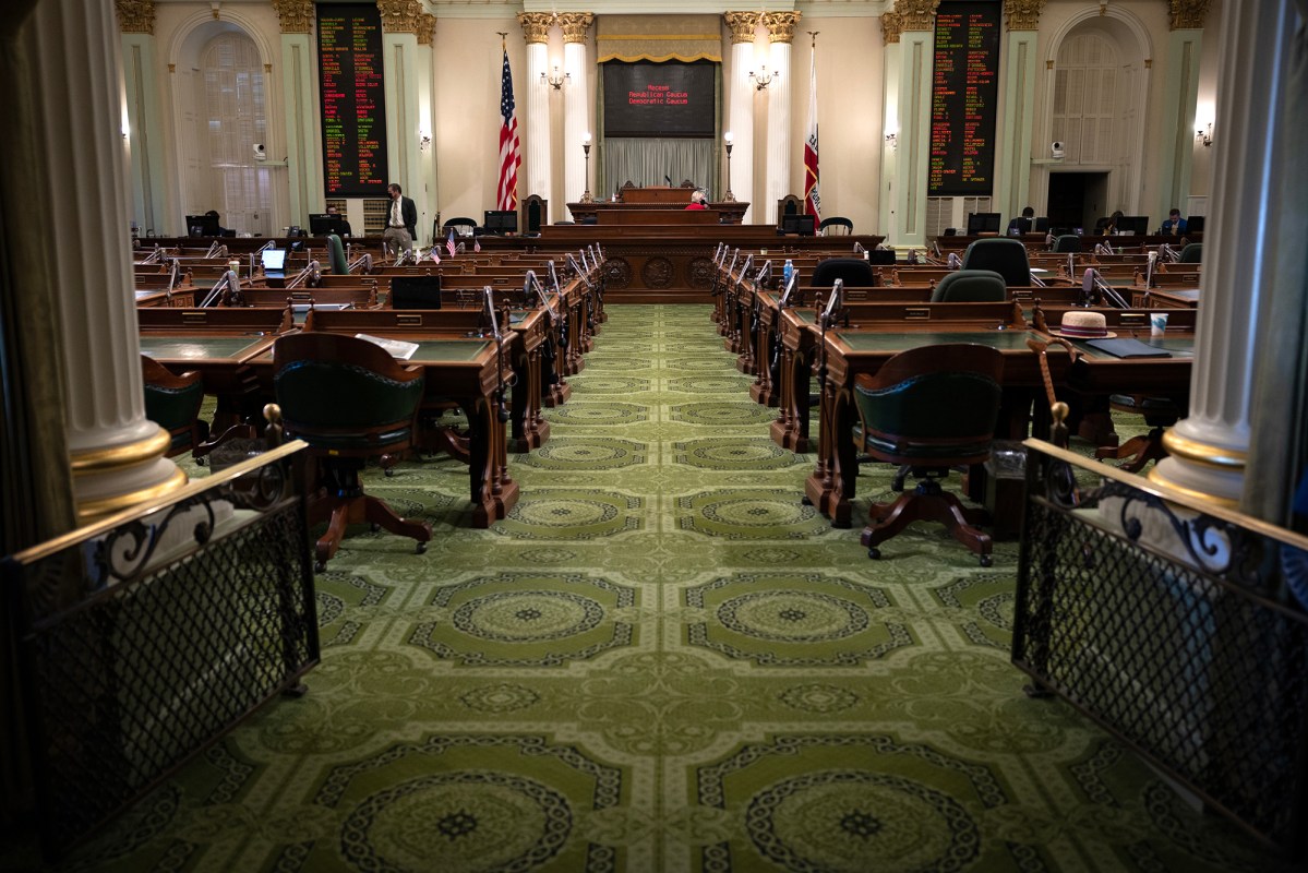 An empty legislative chamber featuring rows of dark wooden desks and green leather chairs arranged neatly on an ornate green carpet. Two large boards with illuminated text hang on either side of the speaker's podium at the front of the room. The American and Californian flags are positioned behind the podium, framed by tall, elegant columns. The quiet setting suggests a pause in legislative activity.