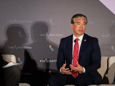 A person in a dark blue suit and a red tie sits in a chair on stage and gestures while they speak. At left, the person's shadow, and that of a moderator are visible on a backdrop with the words "CALMATTERS" emblazoned on it. The setting is a forum or panel.