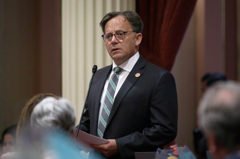 A person dressed in a black suit, white button-up shirt and a black and green tie speaks into a microphone while holding a piece of paper. The heads of other people are visible at the bottom of the frame, as they listen. The setting is a legislative hearing.