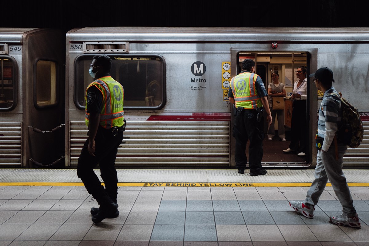 Two workers in yellow and orange safety vests stand on an underground subway platform. One worker speaks to two people on a train, as the doors remain open. Another person walks by and observes the conversation. The train remains stopped at the station.