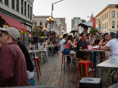 People sit on chairs and tables set up outside a restaurant in a busy area of downtown San Diego.