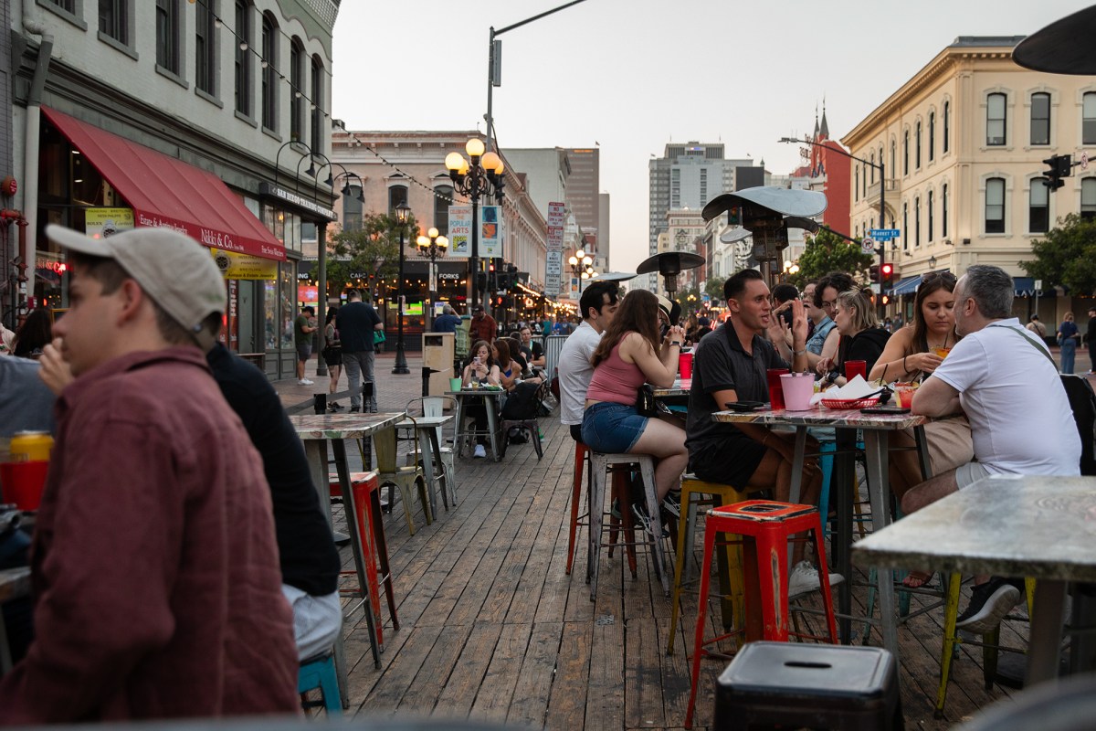 People sit on chairs and tables set up outside a restaurant in a busy area of downtown San Diego.