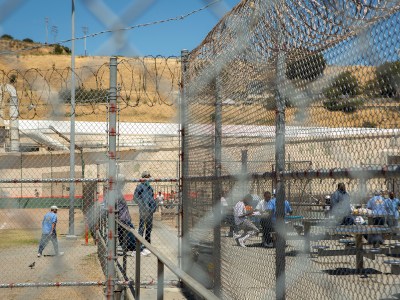 A view through chain-link and barbed wire fences shows an outdoor space within a correctional facility. Several individuals are visible, some standing or walking near the fences and others seated at picnic tables in the background. The scene is set against a backdrop of tan hills, industrial buildings, and a clear blue sky, highlighting the enclosed and institutional environment.