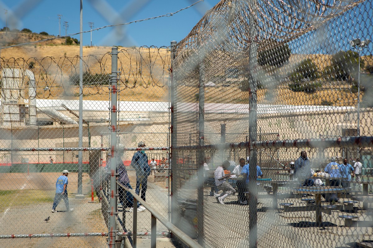 A view through chain-link and barbed wire fences shows an outdoor space within a correctional facility. Several individuals are visible, some standing or walking near the fences and others seated at picnic tables in the background. The scene is set against a backdrop of tan hills, industrial buildings, and a clear blue sky, highlighting the enclosed and institutional environment.