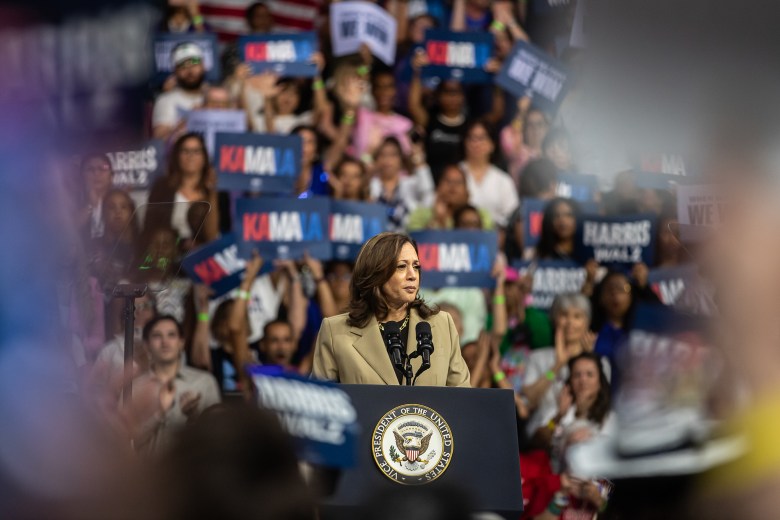 Vice President Kamala Harris, wearing a tan blazer and black shirt, stands behind a podium with as hundreds of supporters stand behind her, waving signs with her name at a campaign rally.