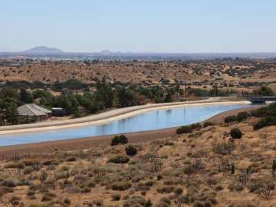 A wide view of an aqueduct, a structure that moves water from one location to another, surrounded by desert shrubs and some housing nearby.