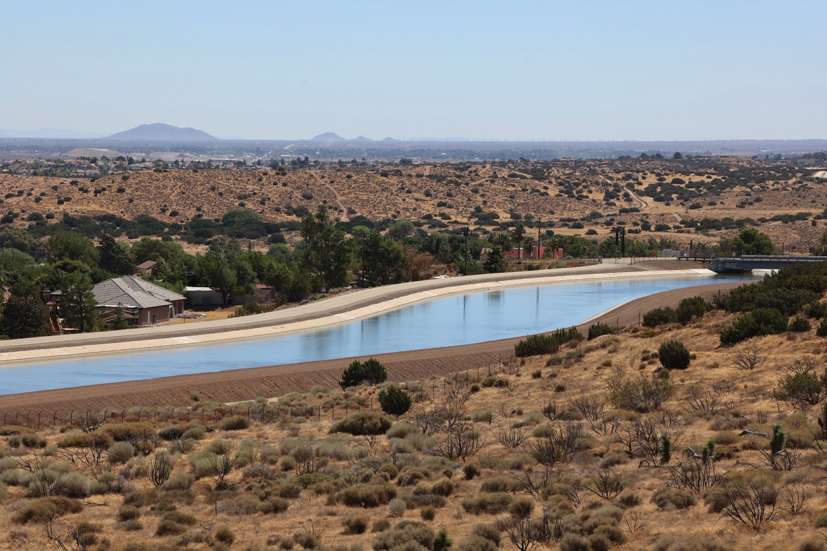 A wide view of an aqueduct, a structure that moves water from one location to another, surrounded by desert shrubs and some housing nearby.