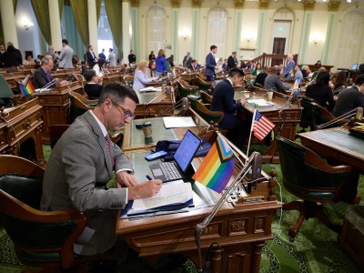 A lawmaker, wearing a gray suit and glasses, sits at their desk on the Assembly floor marking a sheet of paper with a pen.