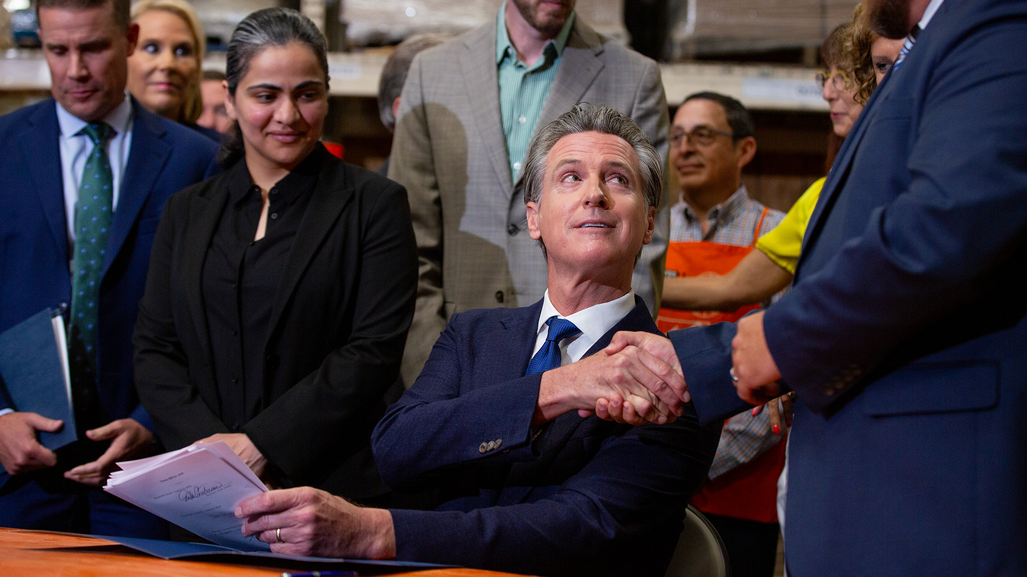 Flanked by state lawmakers, Gov. Gavin Newsom shakes another lawmakers hand in celebration while seated at a desk at a Home Depot in San Jose.