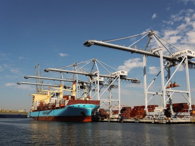 A blue and burnt red cargo ship is unloaded by various cranes at a port. Metal shipping containers are seen in the port. The ship is floating in the water.