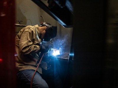 A student in welding mask and other safety gear welds a piece of metal in a work station in a college classroom.