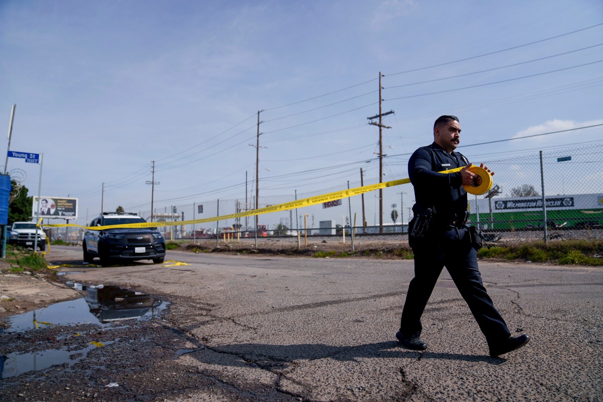 A police officer is unrolling a roll of crime scene tape along an empty road next to a fence and train track to create a crime scene.