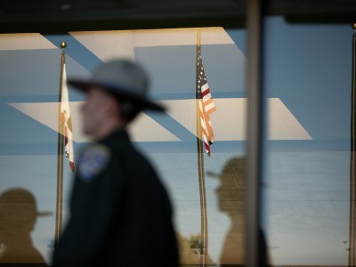 The reflection of a law enforcement officer is seen on a window along with three flag poles with the U.S., California and CHP flags.