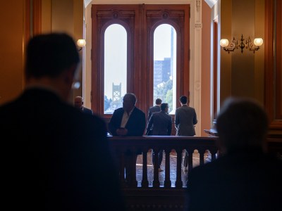 A group of individuals is gathered near large, arched windows in a dimly lit hallway. The silhouettes of a few people stand against the bright backdrop of the windows, which frame a view of a cityscape and a bridge in the distance. The setting appears to be inside a government building, with elegant woodwork and soft lighting from wall-mounted lamps. The scene evokes a sense of quiet anticipation or reflection, with people standing in conversation or gazing out the windows.