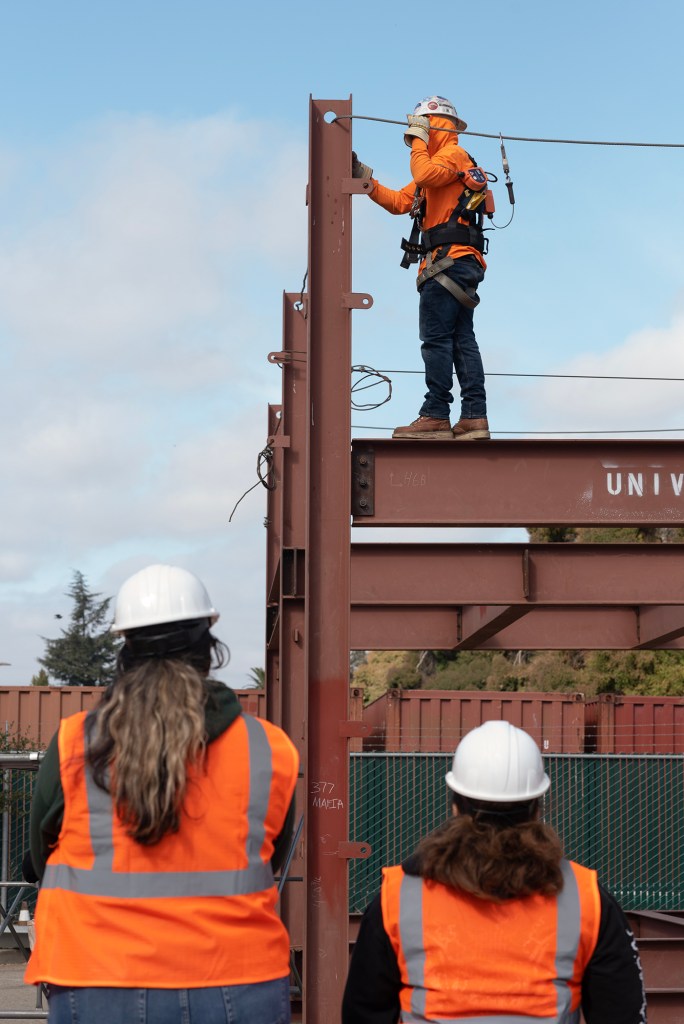 Two people wearing orange safety vests and white safety helmets observe as a person wearing a bright orange sweater and a white safety helmet performs a task on top of a metal structure. The class is part of an iron workers training program.