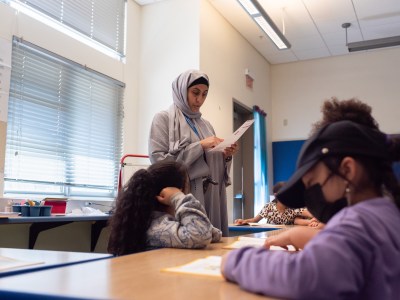 A teacher, wearing a hijab and an abaya, stands nearby a set of desks as she looks over a student's paperwork. Behind her, the blinds of a window are kept closed to keep the room cold during a hot day.