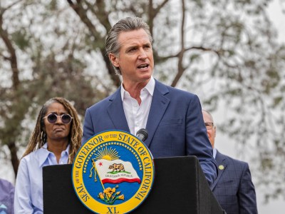 A person, wearing a blue suit and white shirt, stands behind a black podium with the California state seal on it during a press conference.