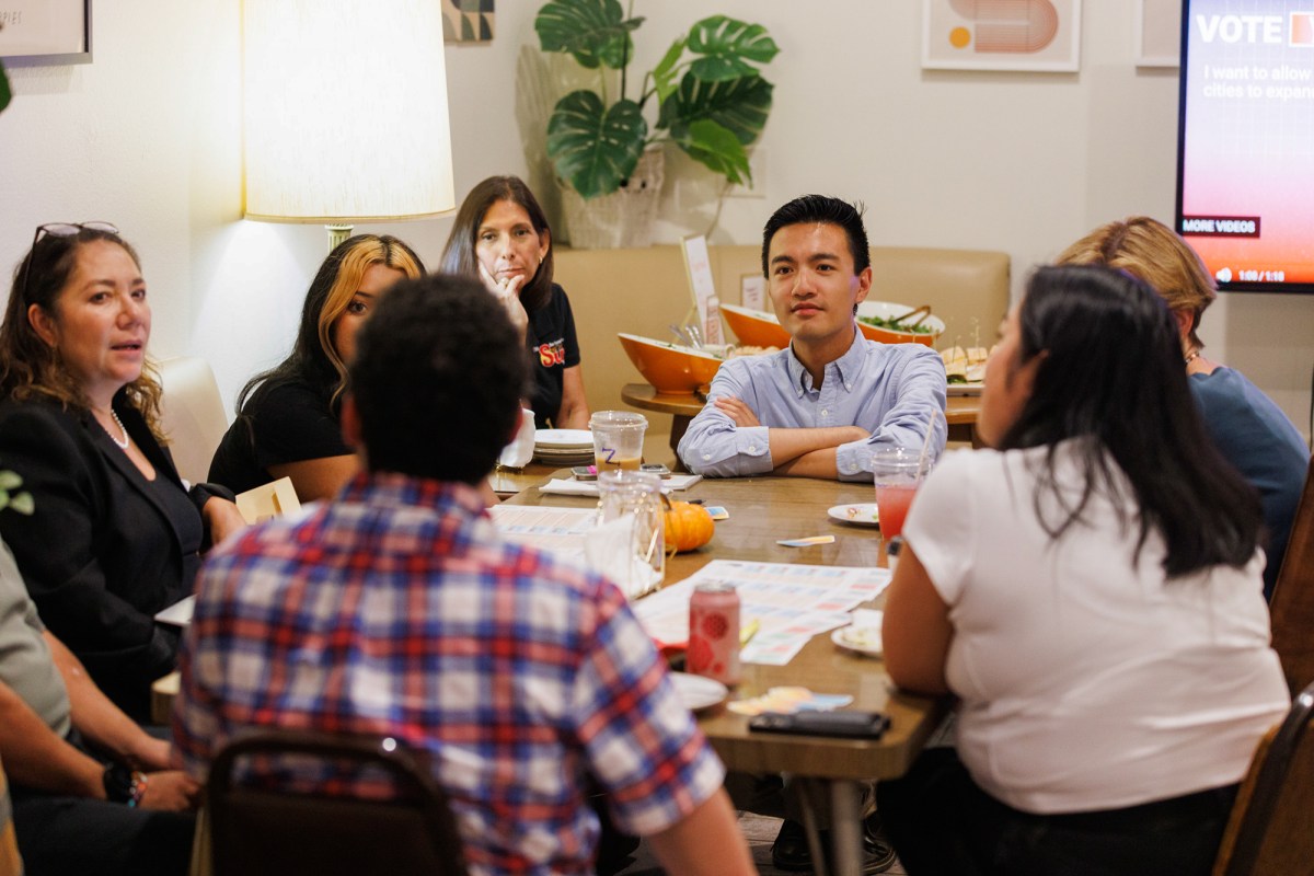 A person, kneeling down with their hands resting on a table, looks at a group of people sitting nearby with pamphlets containing information on ballot propositions at a voter information event.