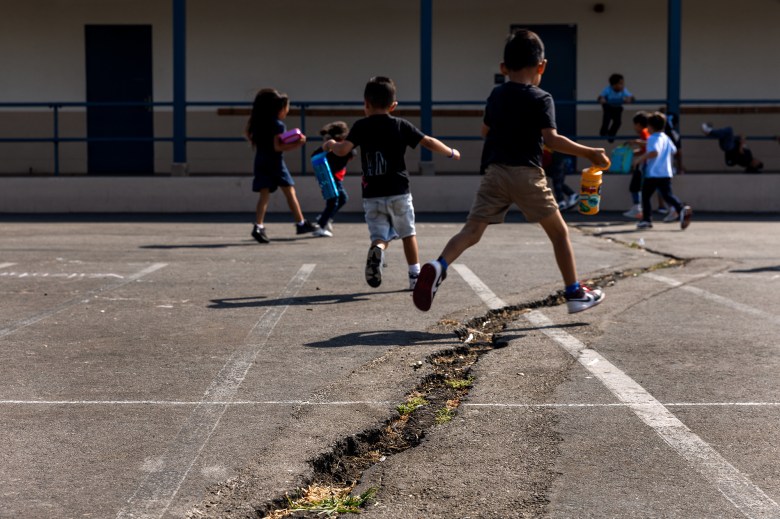 A child jumps over a cracked portion of an asphalt yard at a school. Other children playing can be seen in the background.