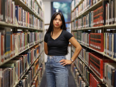 A person stands in the middle of a library aisle, surrounded by shelves filled with books. They are wearing a black V-neck shirt and blue jeans, with one hand on their hip and the other relaxed by their side. The background shows books neatly organized on both sides, leading to a window at the far end.