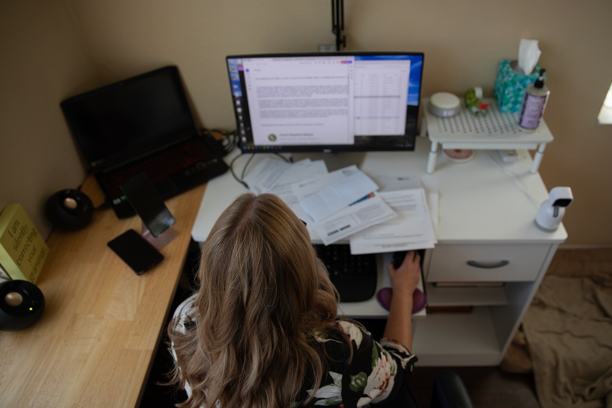 An overhead view of a person with blonde hair sitting in a chair looking at a computer on a desk filled with various papers.