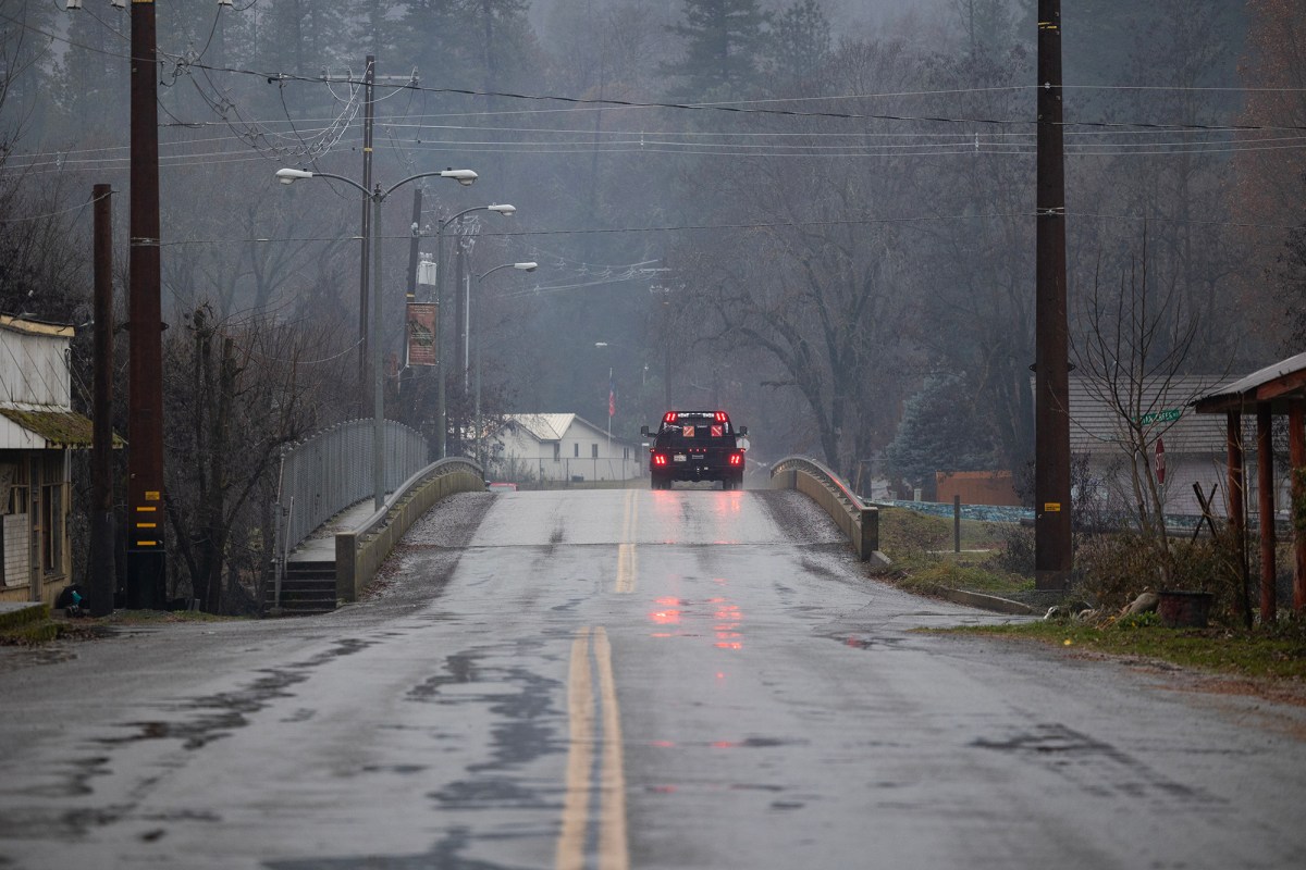 A truck drives over a bridge and down a wet road. The rear of the truck is visible and the brake lights are engaged. The setting is a small, rural, town. The weather is drizzly, foggy and grey.