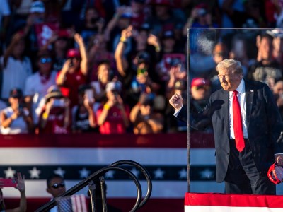 A man in a red tie and suit is gesturing to a crowd while on stage behind bulletproof glass at a political rally.