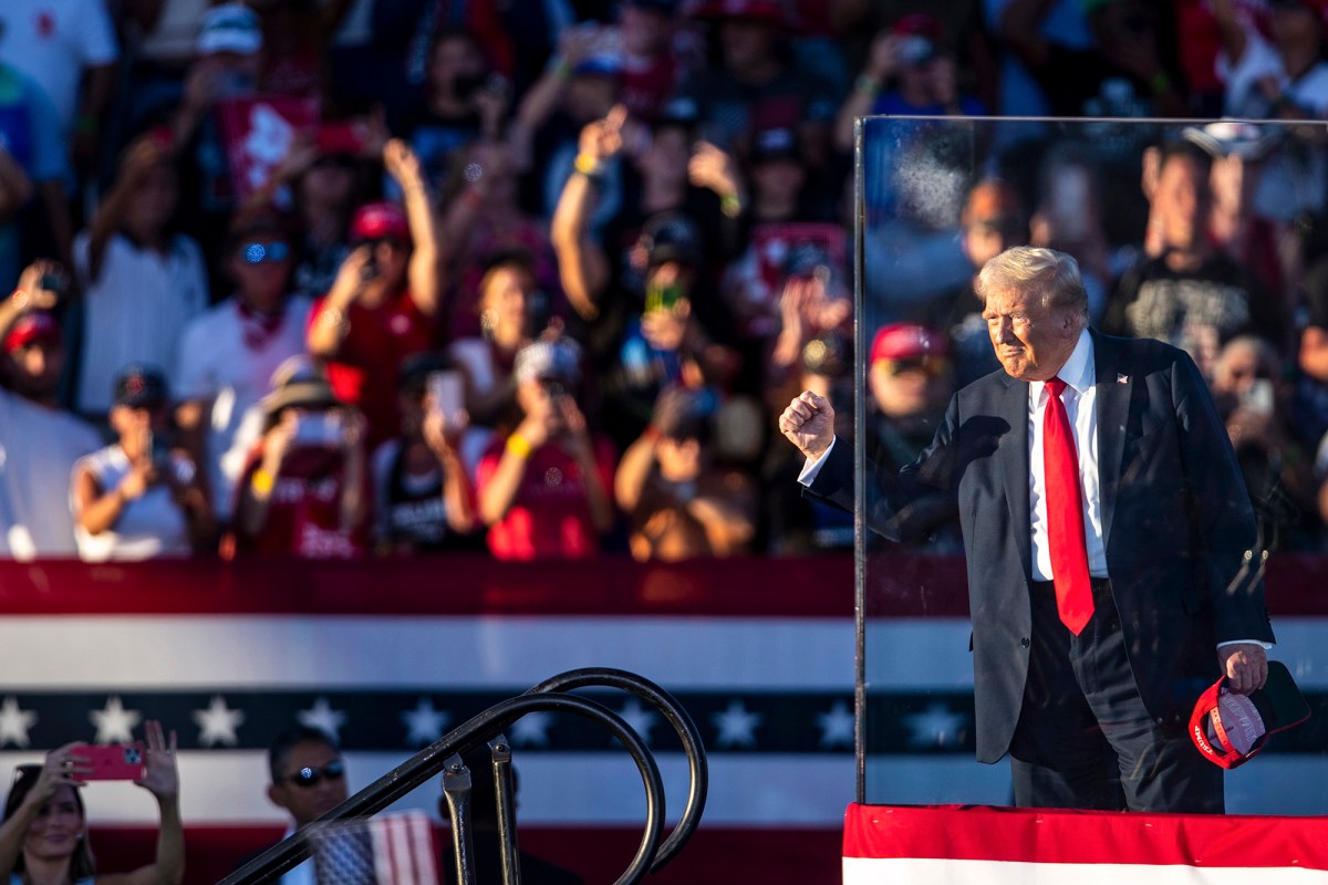 A man in a red tie and suit is gesturing to a crowd while on stage behind bulletproof glass at a political rally.