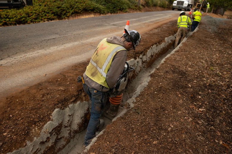 People wearing yellow and orange safety vest and white hard hats work on a trench to lay underground electric cables.