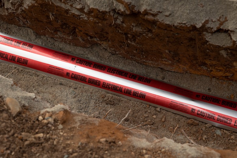 A close-up view of a red and white tap that reads "Caution: Buried Electrical Line Below" inside a trench on a dirt road.