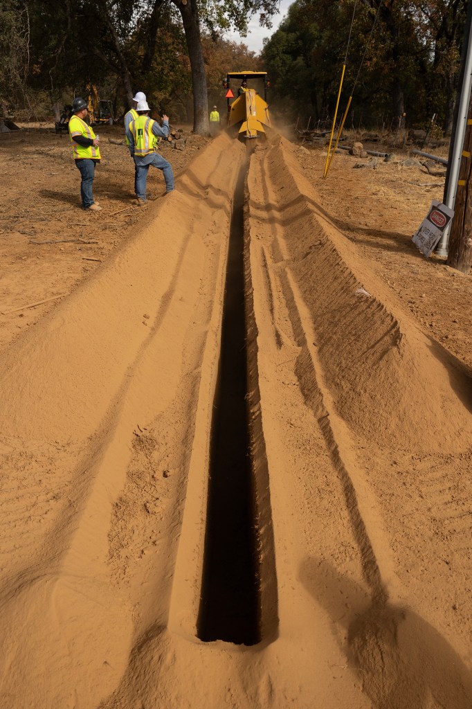 People wearing yellow and orange safety vest and white hard hats work on trench on a dirt road to lay underground electric cables.