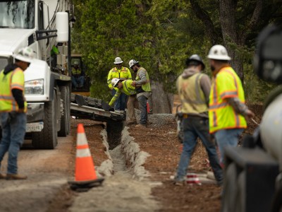 People wearing yellow and orange safety vest and white hard hats work on a trench to lay underground electric cables.