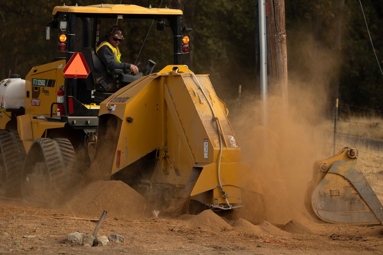 A worker wearing yellow and orange safety vest sits on top a rock saw trencher as they dig a trench where electric cables will eventually be buried.