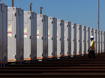 A worker wearing a hard hat and protective clothing walks alongside a row of tall, identical battery storage units under a clear blue sky. The units are aligned precisely on a metal framework, creating a repeating pattern.