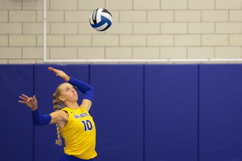 A volleyball player in a yellow jersey with the number 10 prepares to serve, eyes focused on a blue, white, and black volleyball suspended in the air above. The player’s left arm is bent back, ready to swing forward, while the right arm extends outward for balance. The jersey has "San Jose State" written across the chest.