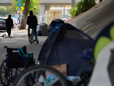 A side view of a man on a bike next to homeless tents lined up along a sidewalk underneath a highway in Sacramento.