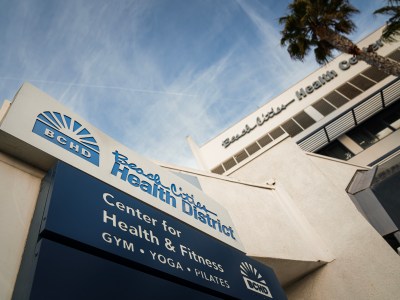 A close-up and angled view of the "Beach Cities Health District" sign at the medical complex in Redondo Beach. The building is visible in the right corner, and the sky above is hazy and blue.