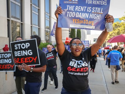 A group of protesters advocating for rent control is gathered outside a building. They hold signs reading "STOP BEING GREEDY" and "THE RENT IS STILL TOO DAMN HIGH!" in bold letters. One person in the foreground wears sunglasses and holds a large sign high above their head, while others around them carry similar signs, showing unity in their demand for affordable housing.