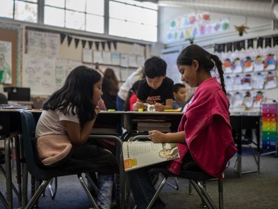 A close-up view of two young students as they sit in chairs facing each other. The student on the right wears a hot pink sweater while looking down at a workbook that rests on her thighs. The student on the left wears a light pink shirt and rest her hands on her chin.