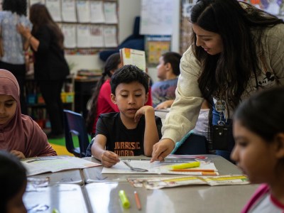 A teacher points at a book while a child sitting at their desk looks at it. Other students are visible in the frame. The setting is a classroom.